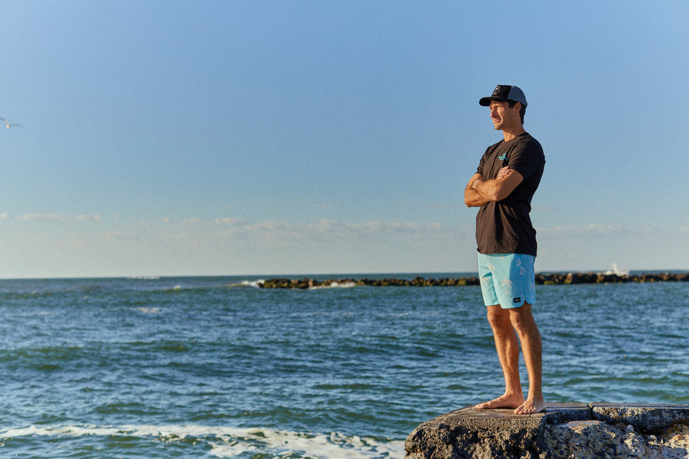 Man staring at ocean in short, tee and hat.
